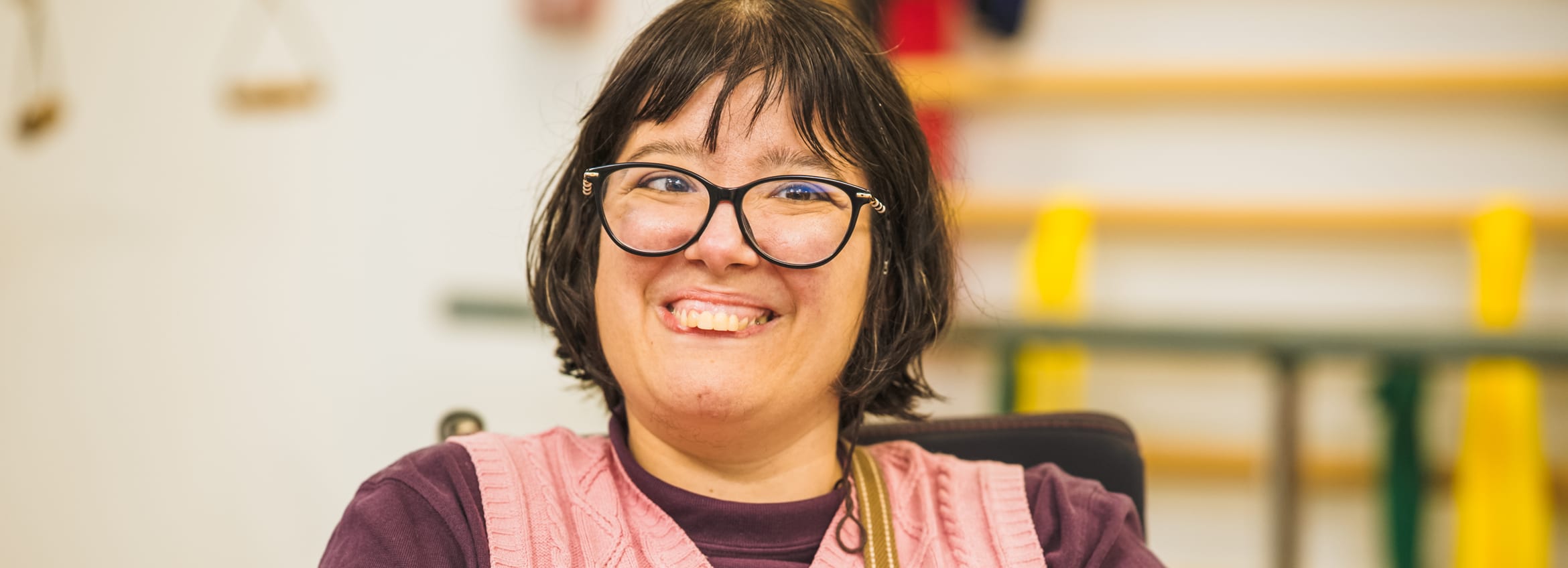 A woman in glasses and a pink knit vest smiles warmly at the camera in a bright community space.