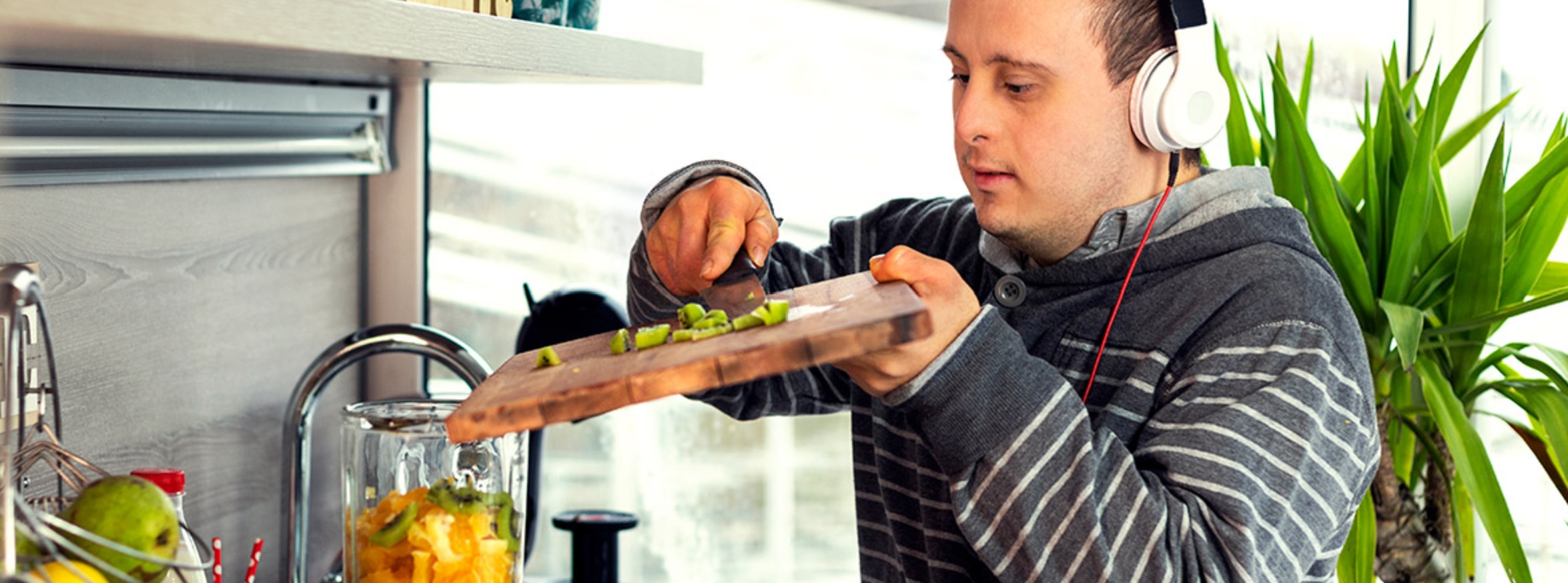 A young man wearing headphones smiles while chopping fresh fruit in a bright kitchen.