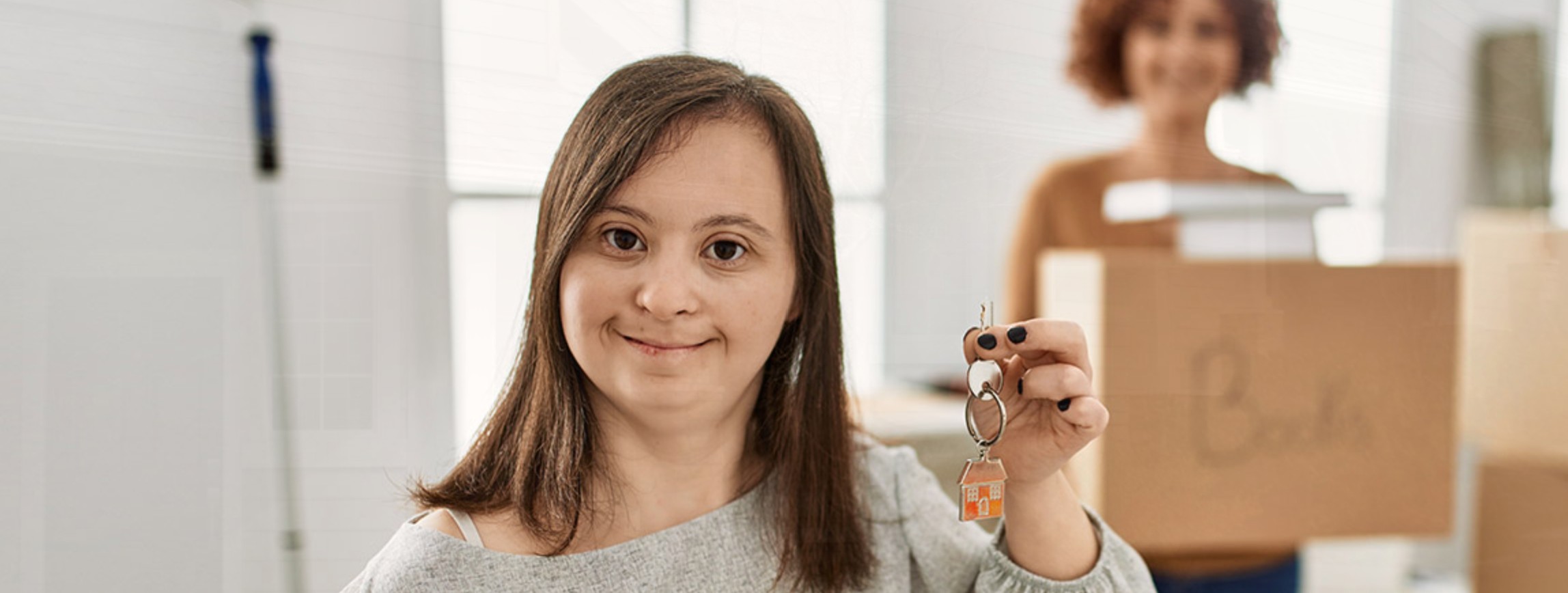 A smiling young woman holds up the keys to her new home on moving day, with a friend carrying a box behind her.
